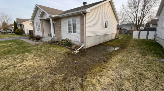 Terrain résidentiel québécois avec zone de sol détrempé et saturé d'eau le long de la fondation en béton d'une maison unifamiliale, illustrant un problème de drainage extérieur